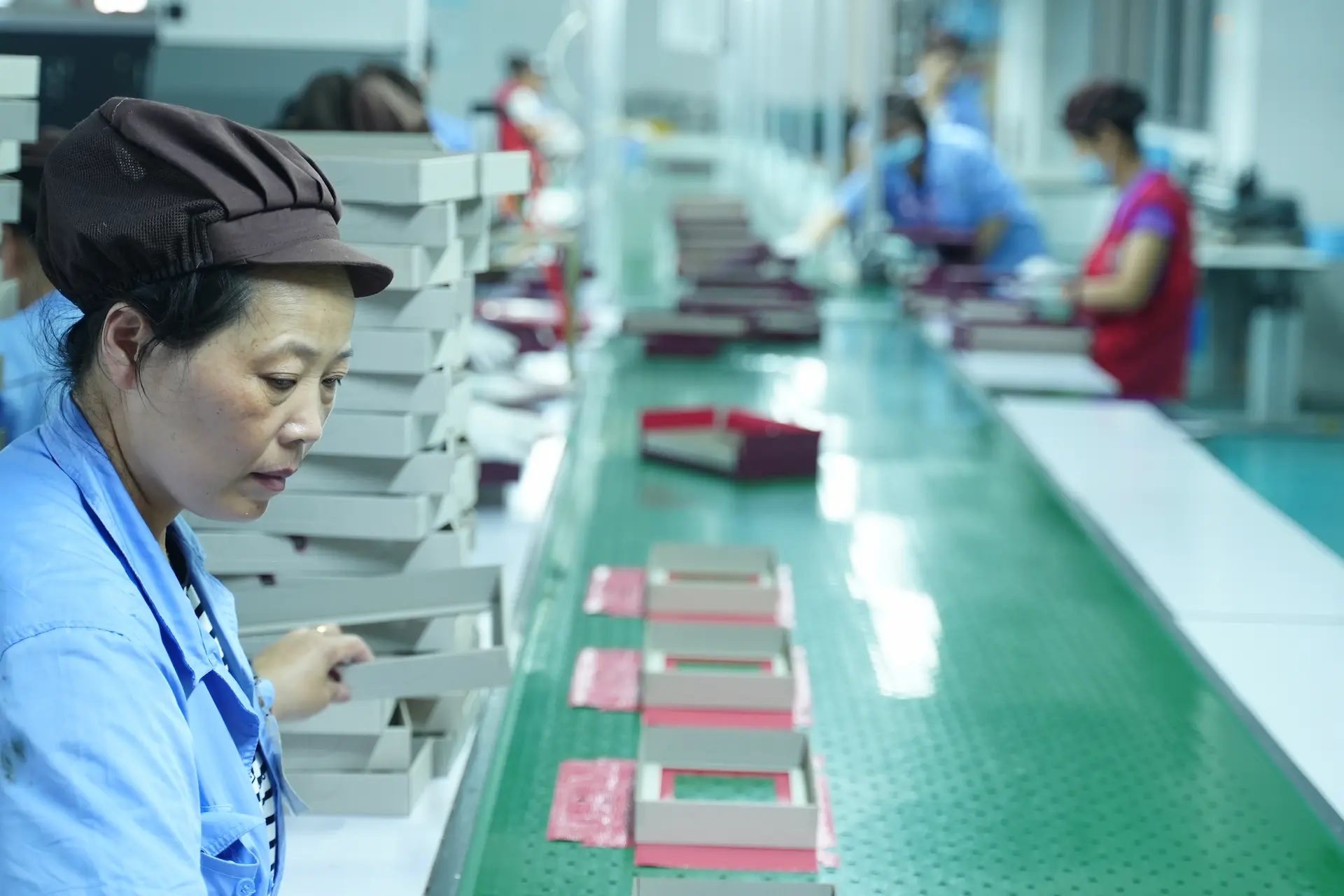 A close-up view of a Witpax employee carefully inspecting paper box components as they move along a green conveyor belt on the factory assembly line.