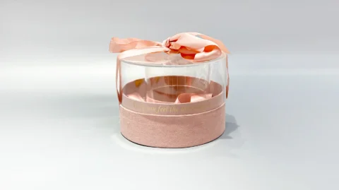 A cylindrical, clear plastic packaging container featuring a decorative pink base and finished with a sophisticated ribbon accent on the lid.