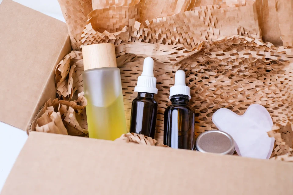 We are a manufacturer of custom box packaging. This image illustrates our product: a cardboard box containing skincare items such as bottles and droppers, securely tucked in crinkled paper for protection. Accompanying these items are a gua sha tool and a compact tin container, all meticulously arranged to showcase the versatility and quality of our packaging solutions, without depicting any branding or text on the packaging itself.