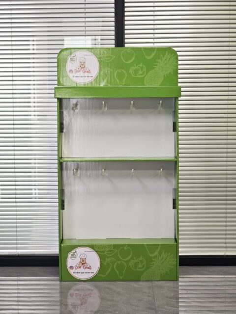 The image shows a green, assembled display box with hanging slots positioned in a room with closed blinds in the background.