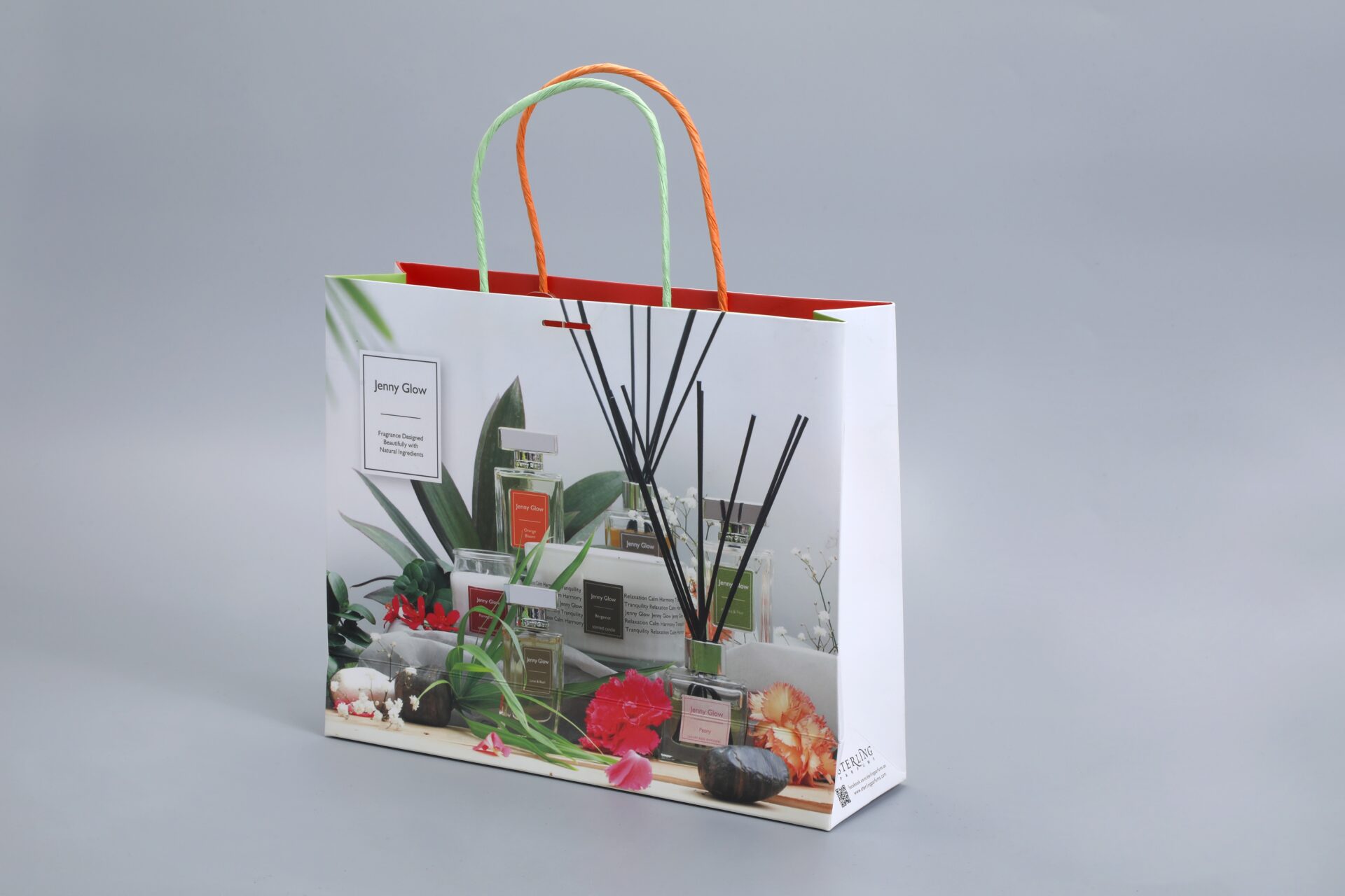 We are a manufacturer of custom box packaging. This image showcases our product: a custom-designed packaging box with sturdy rope handles, featuring a printed design of perfumes, flowers, and reeds on one side.