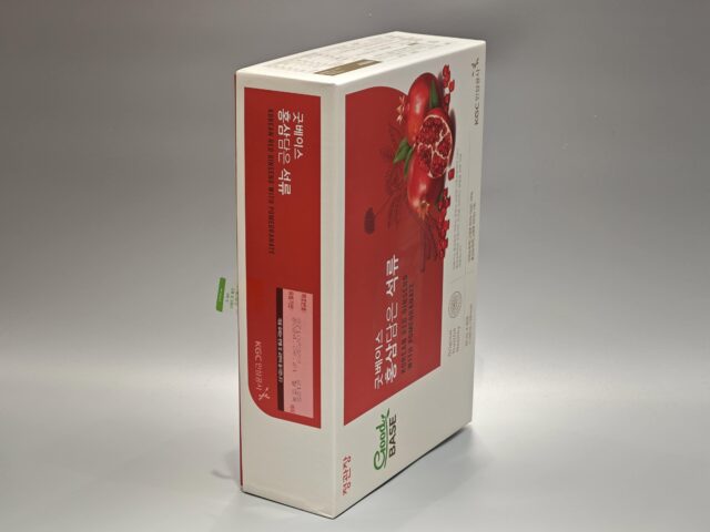 The product is a custom box packaging for a Korean Red Ginseng with Pomegranate product. The box features vibrant and detailed illustrations of ginseng roots and pomegranate fruits, giving it an appealing and eye-catching design. The overall color scheme complements the images, set against a gray surface that enhances the vividness of the artwork. This bespoke packaging highlights both traditional and contemporary aesthetics, reflecting the premium quality of its contents.