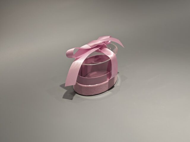 The product is a ellipse, pink custom box with a transparent lid, secured with a pink ribbon